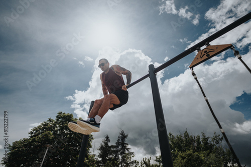 A sporty male with tattoos vigorously trains at an outdoor gym in a park during sunny weather, showcasing fitness and determination.