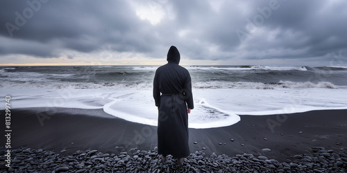 Man in black robes, standing on the shore of a desolate black beach looking over the gray ocean. Dark clouds filly the sky. photo