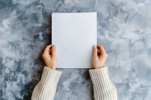 person in a cozy sweater holds a vertical blank paper against a unique marble background, ideal for advertisements or graphic displays. clean, serene setting for custom designs and messages mockup