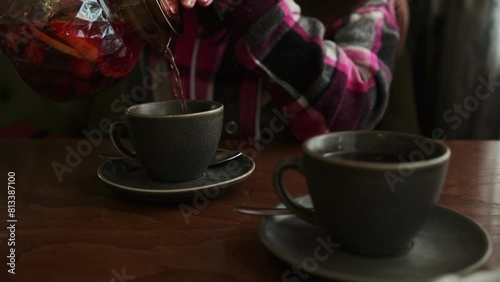 A woman carefully pours hot tea into a cup placed on a wooden table.