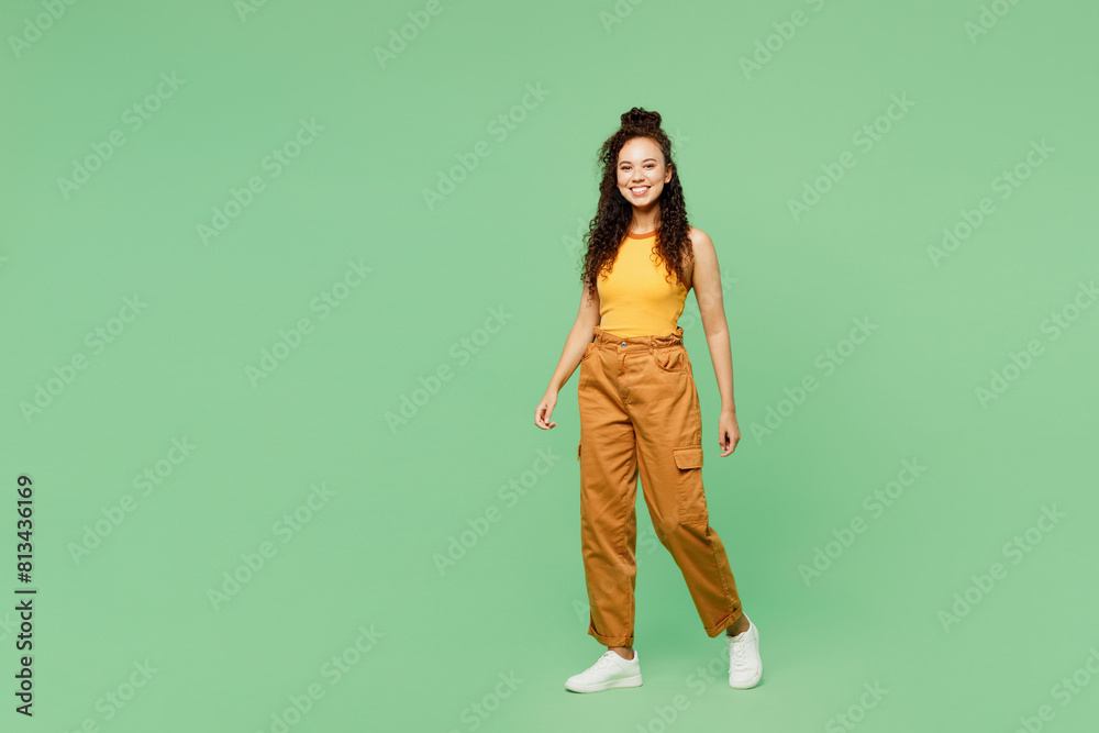 Full body side view young woman of African American ethnicity wear yellow tank shirt top walking going look camera isolated on plain pastel light green background studio portrait. Lifestyle concept. Full body side view young woman of African American ethnicity wear yellow tank shirt top walking going look camera isolated on plain pastel light green background studio portrait. Lifestyle concept.