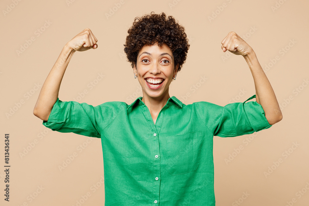 Young strong sporty woman of African American ethnicity wear green shirt casual clothes showing biceps muscles on hand demonstrate strength power isolated on plain beige background. Lifestyle concept. Young strong sporty woman of African American ethnicity wear green shirt casual clothes showing biceps muscles on hand demonstrate strength power isolated on plain beige background. Lifestyle concept.