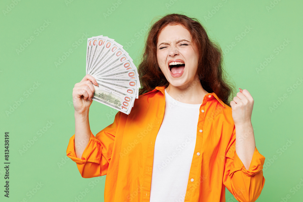 Young ginger woman she wear orange shirt white t-shirt casual clothes hold in hand fan of cash money in dollar banknotes do winner gesture isolated on plain pastel green background. Lifestyle concept. Young ginger woman she wear orange shirt white t-shirt casual clothes hold in hand fan of cash money in dollar banknotes do winner gesture isolated on plain pastel green background. Lifestyle concept.