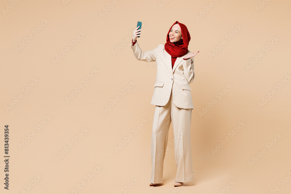 Full body young Arabian Asian Muslim woman wears red abaya hijab suit clothes doing selfie shot on mobile cell phone waving hand isolated on plain beige background. UAE middle eastern Islam concept. Full body young Arabian Asian Muslim woman wears red abaya hijab suit clothes doing selfie shot on mobile cell phone waving hand isolated on plain beige background. UAE middle eastern Islam concept.