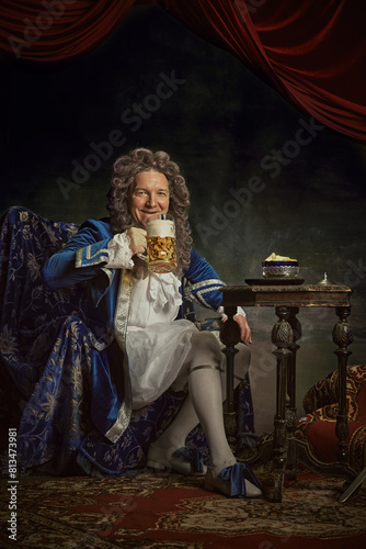 Happy elderly man  dresses as medieval person  king drinks alcohol drink  beer against vintage studio background. Concept of comparisons of eras  fusion of modernity and history  Oktoberfest. Ad
