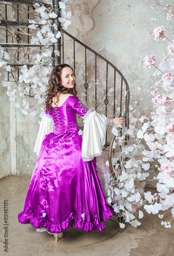 Happy beautiful woman in fantasy white and purple rococo style medieval dress turning standing near the stairway with white flowers