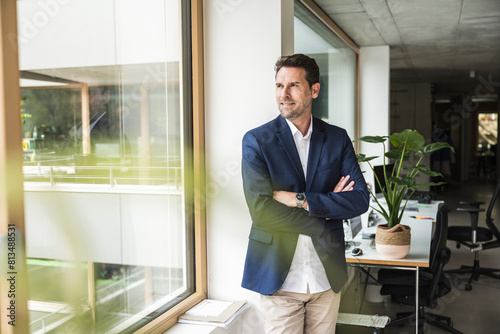 Mature businessman standing with arms crossed looking out through window of office photo