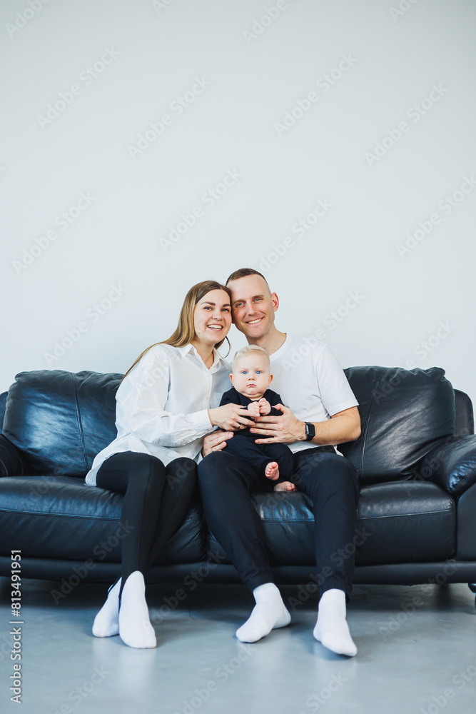 A young married couple with a small child is photographed sitting on a sofa on a white background. Caring parents look at their baby while holding it in their arms. A young married couple with a small child is photographed sitting on a sofa on a white background. Caring parents look at their baby while holding it in their arms.