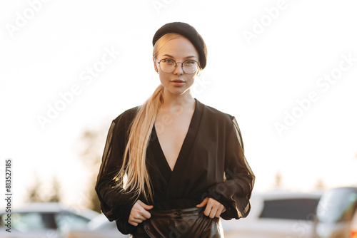 Cute young girl with light daytime make-up and bun hair, with clear glasses on her eyes and a black beret on her head, wearing a black blouse and black leather pants, standing on a blurred background