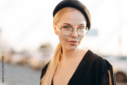 Portrait of a flirty girl with light daytime makeup, clear glasses on her eyes and a black beret, wearing a black blouse on a blurred background