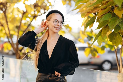 Portrait of a sophisticated girl with light daytime make-up and bun hair, with clear glasses on her eyes and plack leather pants, stands against a background of yellow-green leaves on a tree
