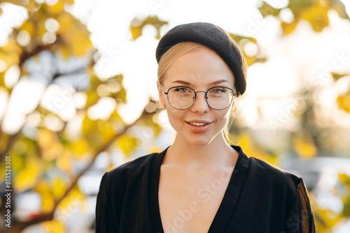 The face of a beautiful girl with light daytime makeup, transparent glasses on her eyes and a black beret, wearing a black blouse against the background of a parking and autumn leaves on a tree