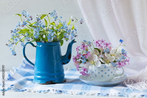 Still life with spring flowers forget-me-nots and others in a cup and jug on the white table.