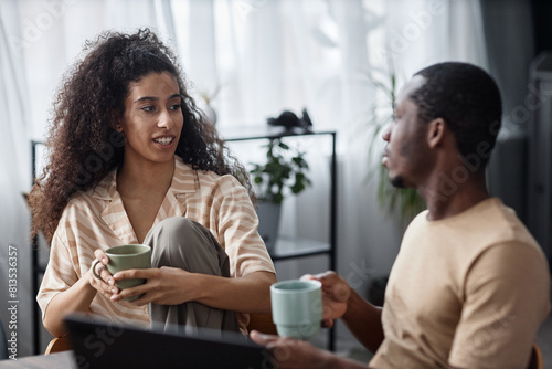 Young biracial woman and her African American boyfriend holding cups with tea or coffee sitting in living room and talking