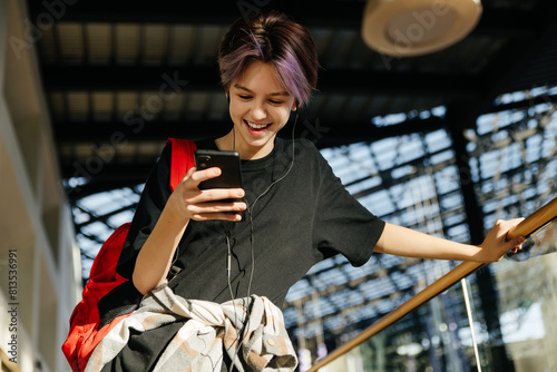 A teenage girl with short hair in a shopping mall, looking at the phone and smiling, headphones in her ears.