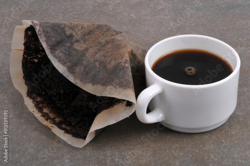 Filtre en papier utilisé rempli de marc de café avec une tasse de café sur fond gris
