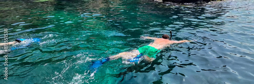 Two people swimming with snorkels in clear turquoise waters near rocky cliffs, implying summer vacations and water activities