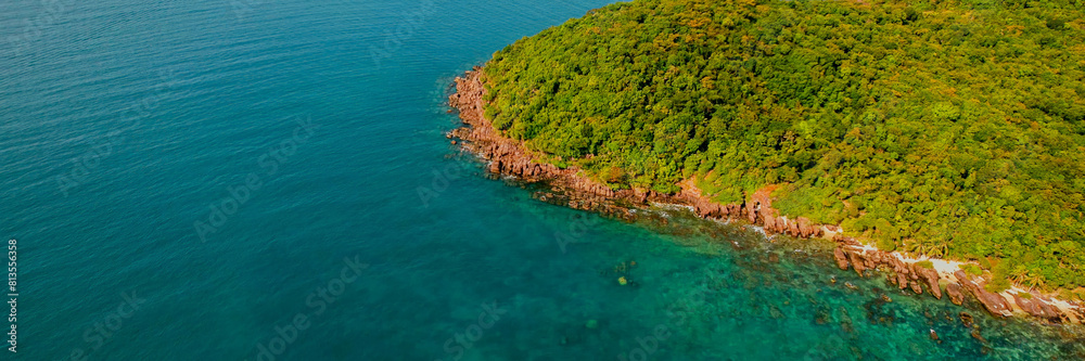 Aerial view of a rugged coastline with red rock formations and lush greenery meeting the turquoise sea, suitable for Earth Day and nature conservation topics