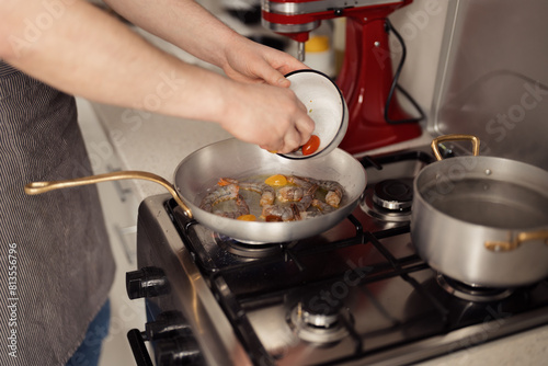 Person cooking, adding cherry tomatoes to a frying pan with shrimps on a stove.