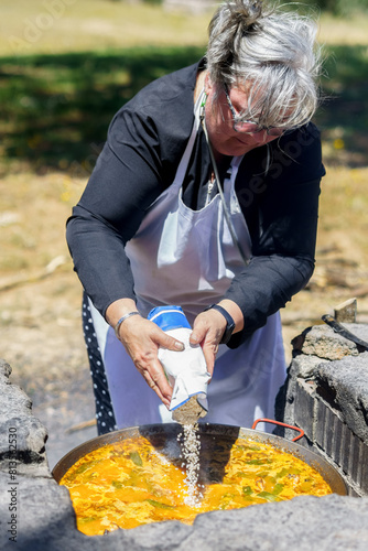 woman cook in white apron pouring rice into a typical Spanish paella cooked in the countryside on the fire.