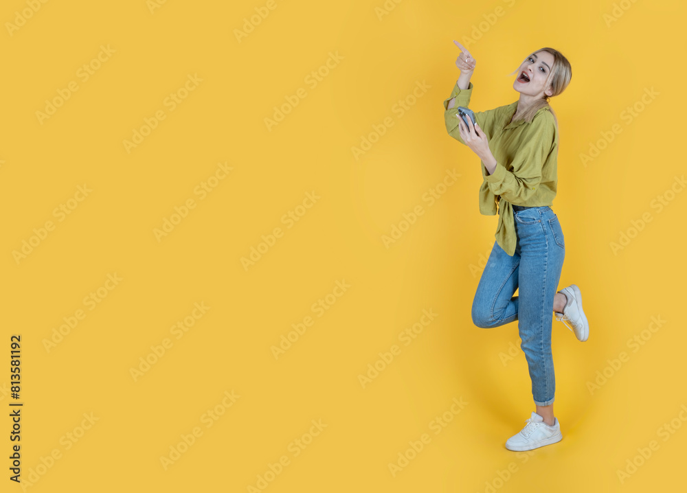 Fototapeta premium Pointing copy space, full body side view young caucasian fun excited happy overjoy blonde woman pointing copy space hold use mobile phone or smartphone yellow studio background copy space banner idea.