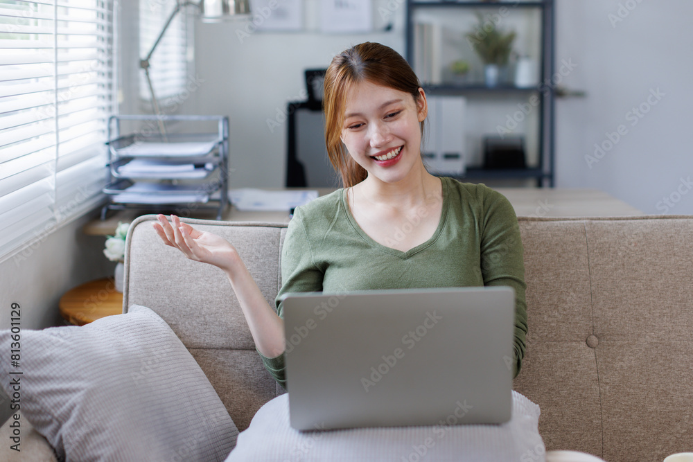 Naklejka premium Attractive happy young girl business or student studying at the college library, sitting at the desk, using laptop computer, having video chat, waving