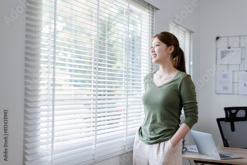  Young Asian business woman smiling confidently while standing alone in a bright modern office