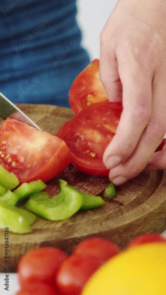 The girl slices the red tomato for salad on the kitchen board with vegetables for vegetarian food. Vertical video. The girl slices the red tomato for salad on the kitchen board with vegetables for vegetarian food. Vertical video.