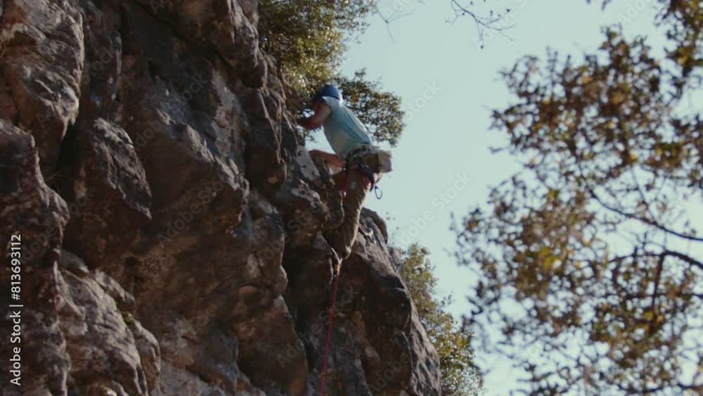 male climber, wearing a casual blue shirt and tan pants, is actively engaged in scaling a rugged cliff. The image captures his focused exertion as he uses both hands to secure his grip on the uneven male climber, wearing a casual blue shirt and tan pants, is actively engaged in scaling a rugged cliff. The image captures his focused exertion as he uses both hands to secure his grip on the uneven