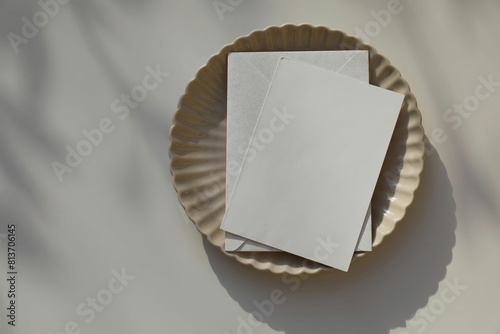 Blank greeting card, invitation mockup, envelope in sunlight. Scalloped ceramic plate on beige table background. Soft shadows. Neutral wedding stationery. Food, restaurant, craft concept, flat lay