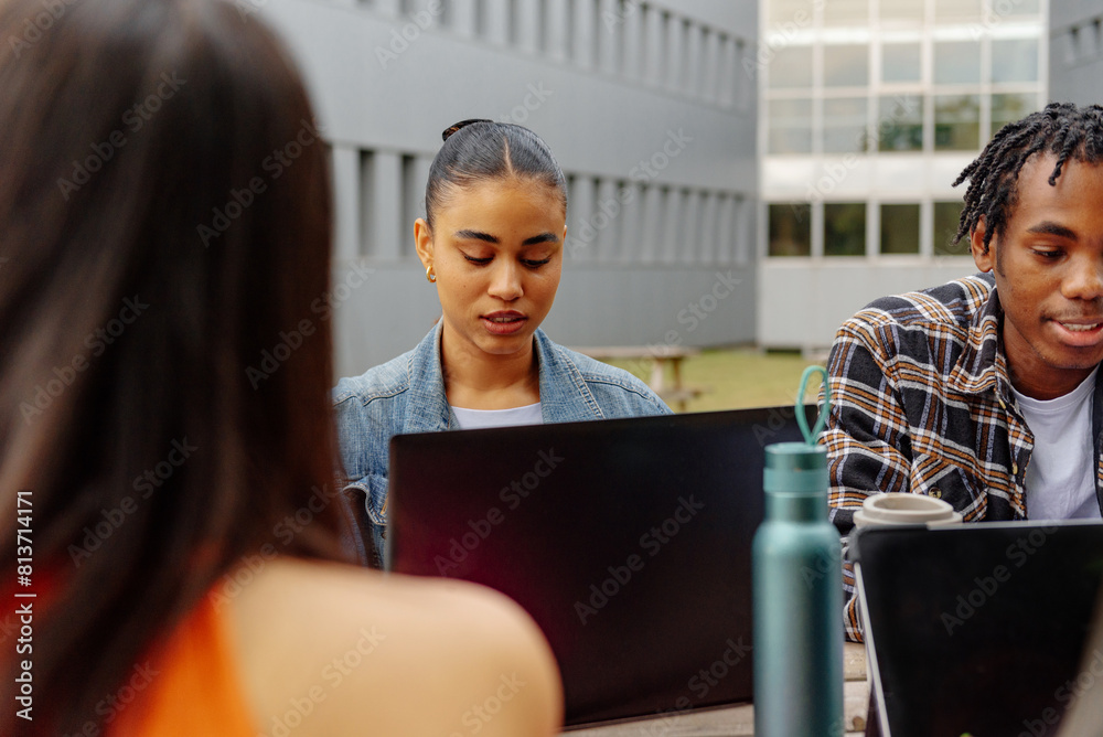 Latina female student sitting on a campus bench using a laptop computer while doing classwork with her classmates. Latina female student sitting on a campus bench using a laptop computer while doing classwork with her classmates.