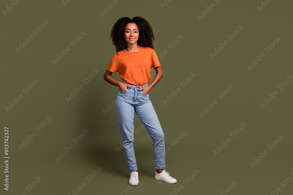 Full size photo of confident adorable girl wear orange t-shirt denim pants holding palms in pockets isolated on dark khaki background