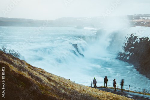 Fototapeta Naklejka Na Ścianę i Meble -  Tourist visit stand on viewpoint Gullfoss waterfall in Iceland, Cinematic beautiful majestic winter waterfall cover by snow and ice. Slow motion. Purple Sunset sky