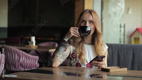 A young woman in casual attire sits by the window in a cafe, drinking coffee. Looking at the phone, typing text on social networks.