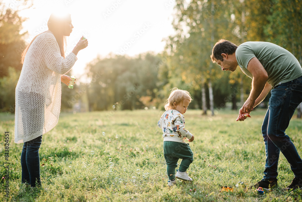 Fototapeta premium Mother blows soap bubbles. Toddler little cute son, dad walking and playing in spring garden. Family catches soap bubbles on green grass in park enjoying sunny summer day. Outdoors creative activities