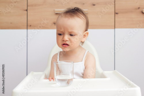 Unhappy baby sitting in a baby chair in the kitchen. Child drinks water. Feeding and childhood