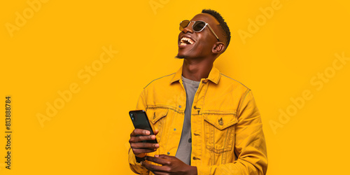 Happy African millennial man laughing with mobile phone on yellow studio backdrop. Joyful black man wearing jeans jacket, sunglasses smiling looking aside. Great new mobile App. People and gadgets