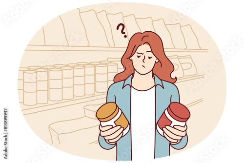 Doubting woman standing in grocery store with two similar cans and reading composition before buying. Girl has doubts buying honey or peanut butter and needs help of consultant working in supermarket