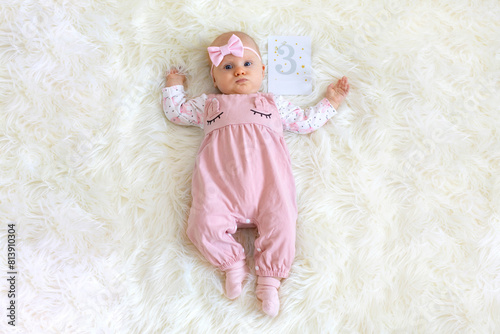 A baby girl, wearing a pink onesie and a bow on her head, lies on a soft, white blanket with a card beside her indicating she is three months old. She looks up with curious eyes, seemingly enjoying he