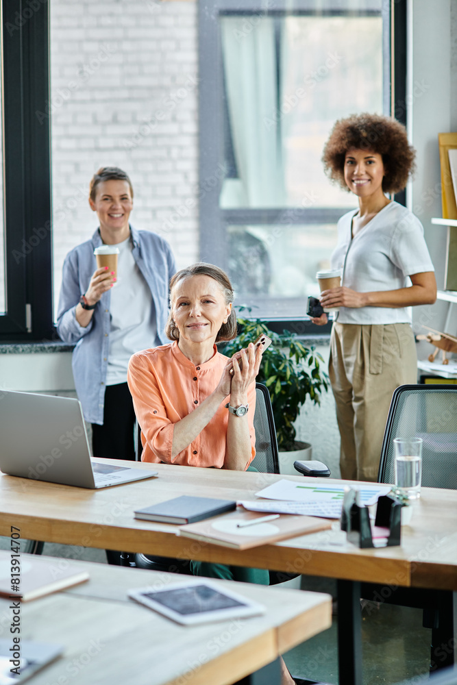 Joyous businesswoman talking on phone while using laptop, with her diverse colleagues on backdrop.