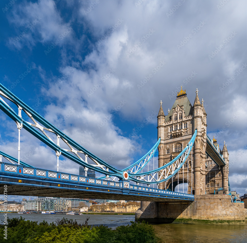Fototapeta premium View from the edge of the River Thames of London's Tower Bridge illuminated by the morning sun under a blue sky with white clouds and ferry traffic on the river and docks in the background. United Kin