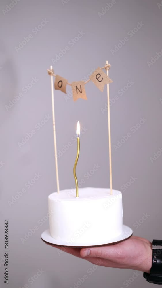 Male hand holding a white cake with a sign of one year celebration. A candle burning on the dessert for first baby birthday. White backdrop. Vertical video. Male hand holding a white cake with a sign of one year celebration. A candle burning on the dessert for first baby birthday. White backdrop. Vertical video.