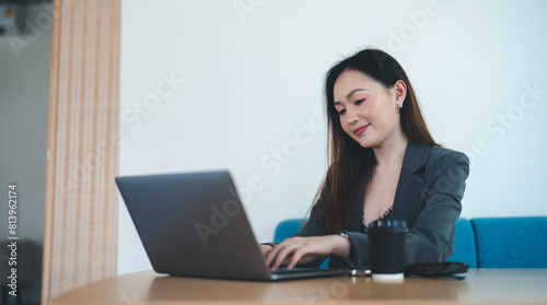 Asian young beautiful happy professional successful businesswoman designer sitting smiling at workstation desk using laptop notebook computer and smartphone working remotely online at home office