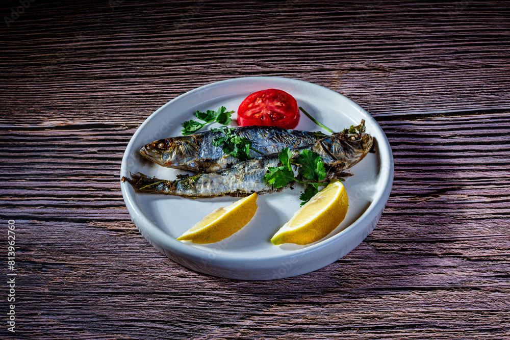 A dish with two oven-roasted sardines, accompanied with lemon, parsley and tomato. A dish with two oven-roasted sardines, accompanied with lemon, parsley and tomato.