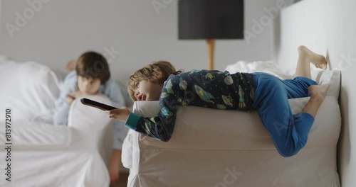 Two kids lounge idly in a living room, one lying on a couch with a remote in hand, eyes glued to the TV, the other sitting nearby, a picture of lethargy.