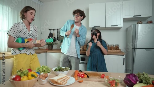 Caucasian skilled father, mother and asian daughter making breakfast while dancing together. Skilled mom playing ukulele while preparing vegetable at modern kitchen. Healthy food concept. Pedagogy.