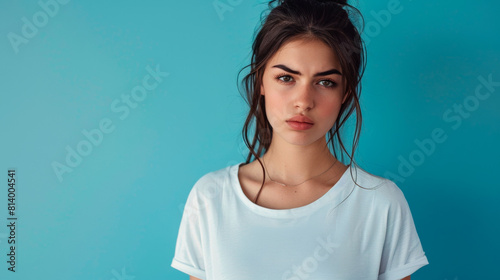 Portrait of an attractive young sad and unhappy brunette woman on a blue background with a copy space. A sweet, upset and offended girl in a simple white T-shirt. photo