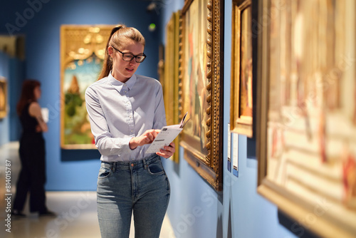 Thoughtful young Caucasian woman wearing glasses and looking at exhibition. Concept of Museum Day.
