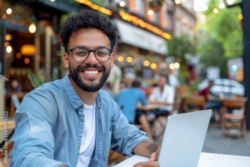 A friendly young man with a bright smile and curly hair productively works on his laptop at a vibrant cafe, enjoying the lively atmosphere photo