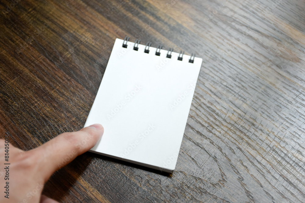 Obraz premium Hand pointing at blank white paper of notebook on wooden patterned desk background at office.